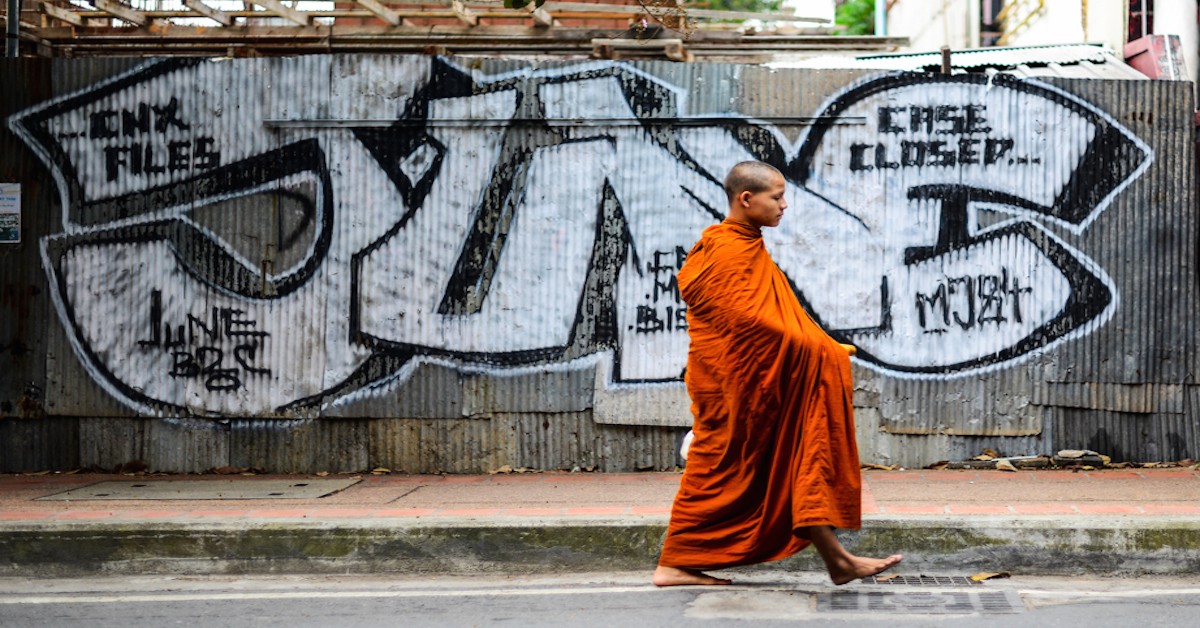 Monk in Orange Robe Walks Barefoot on Urban Street in Front of Graffiti