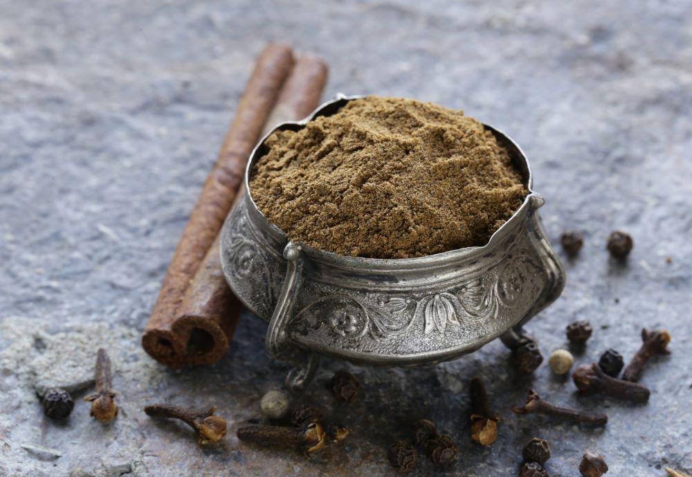 Metal Bowl of Garam Masala Powder, next to Cinnamon Sticks, Cloves and Black Peppercorns.